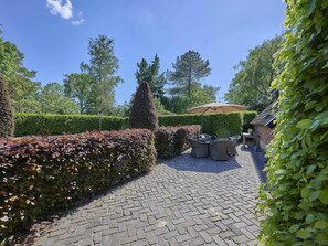 Ein gepflasterter Patio-Bereich mit Tisch und Stühlen unter einem großen Sonnenschirm, umgeben von gepflegten Hecken und Bäumen.