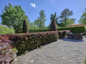 A paved outdoor area with a hedge, a table with chairs, and a large umbrella.
