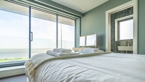 A large bed with a view of the ocean, a bathroom with a mirror, and a window with a view of the ocean. - Whitstable | 4 bd | Pool | Games Room | Cinema (Kent)