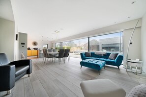 A modern living room with a blue sofa, a dining area with a white table and chairs, and large windows offering a view of the outside. - Whitstable | 4 bd | Pool | Games Room | Cinema (Kent)