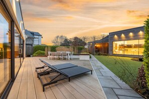 A modern outdoor patio with a dining table and chairs, a wooden deck, and a well-maintained lawn. - Whitstable | 4 bd | Pool | Games Room | Cinema (Kent)