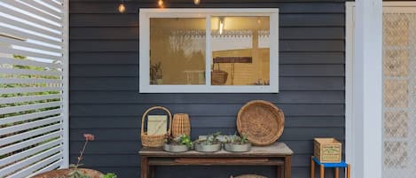 Un espace extérieur abrité, avec une table où sont exposés des paniers et des plantes en pot, une brouette remplie de plantes et une fenêtre décorée de guirlandes lumineuses.