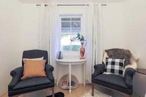 Ein Eckzimmer mit zwei Sesseln, einem kleinen Tisch mit einer Vase und einem Fenster mit Vorhängen.