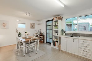Eine helle Küche mit weißen Schränken, ein Essbereich mit Holztisch und -stühlen sowie ein großes Fenster mit Blick auf einen Garten.