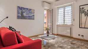A living room with a red sofa, a glass coffee table with a vase, a painting of a night sky, and a window with blinds. - Quadri D'oro (Loano)