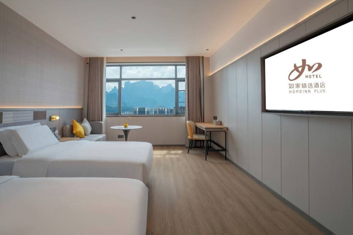 A hotel room with two beds, a small table, a chair, and a large window with a view of mountains.