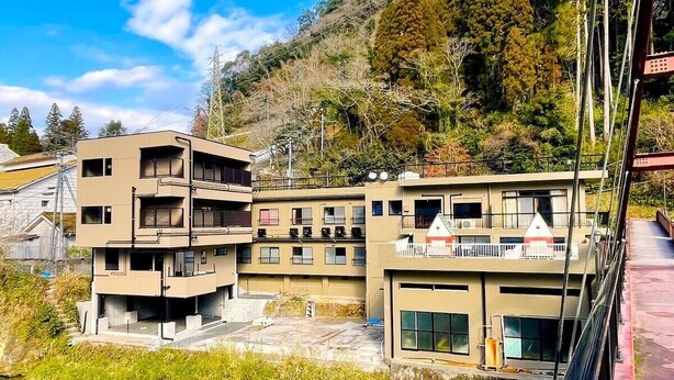 A modern multi-story building with balconies, located near a body of water and surrounded by greenery. - Myoukenkan (Kirishima)