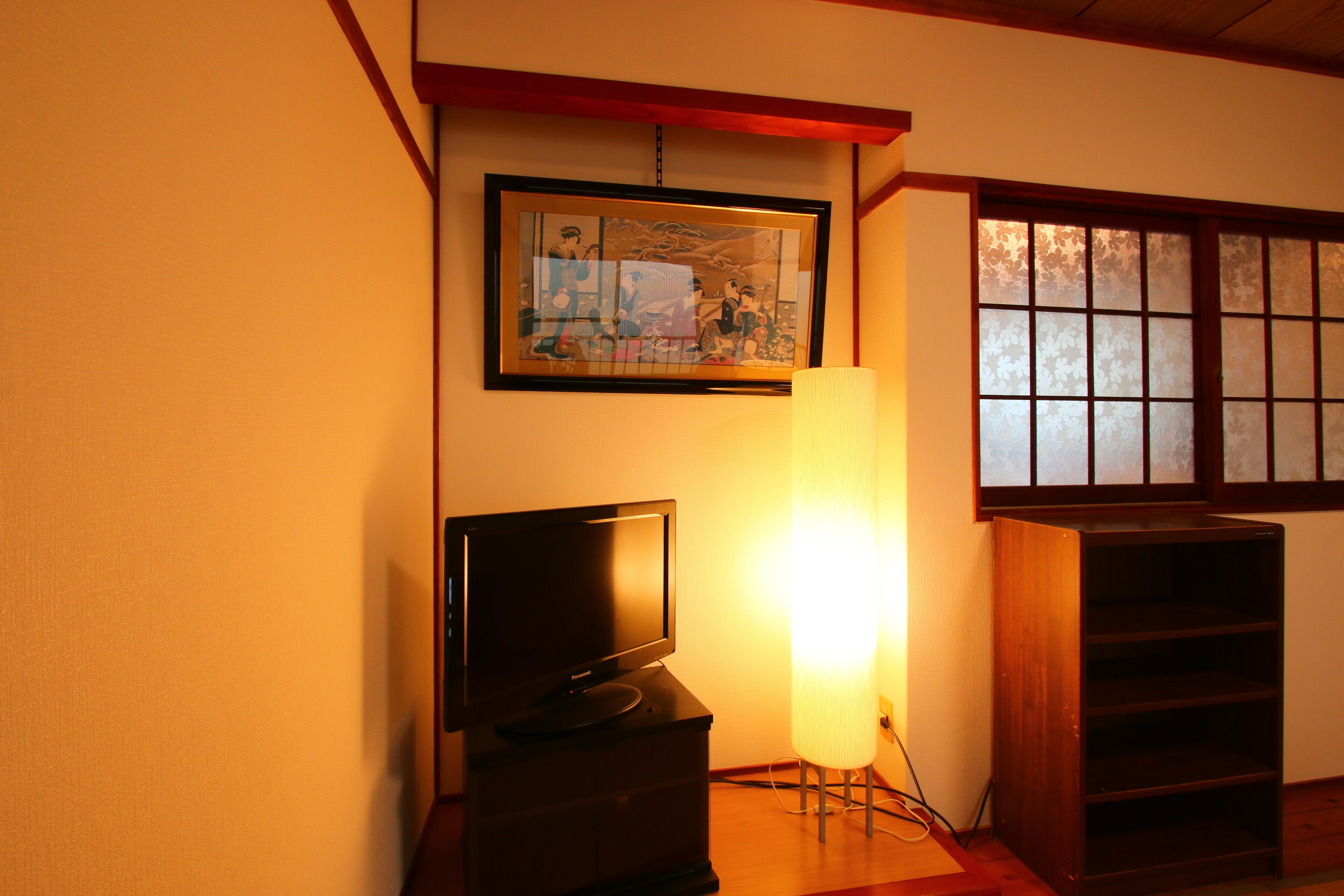 Una habitación con un televisor sobre un soporte de madera, una obra de arte enmarcada en la pared, una lámpara de pie y una ventana tradicional con marcos de madera.