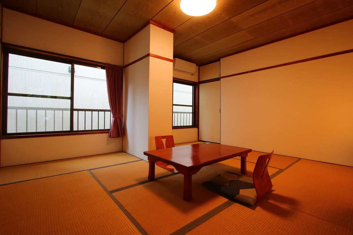 Une pièce de style traditionnel avec des tatamis, une table et des chaises en bois, de grandes fenêtres et un ventilateur de plafond.