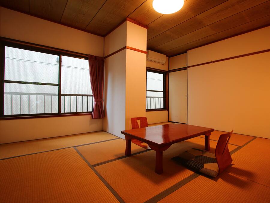 Um quarto tradicional em tatame com uma mesa e cadeiras de madeira, janelas amplas e um ventilador de teto.