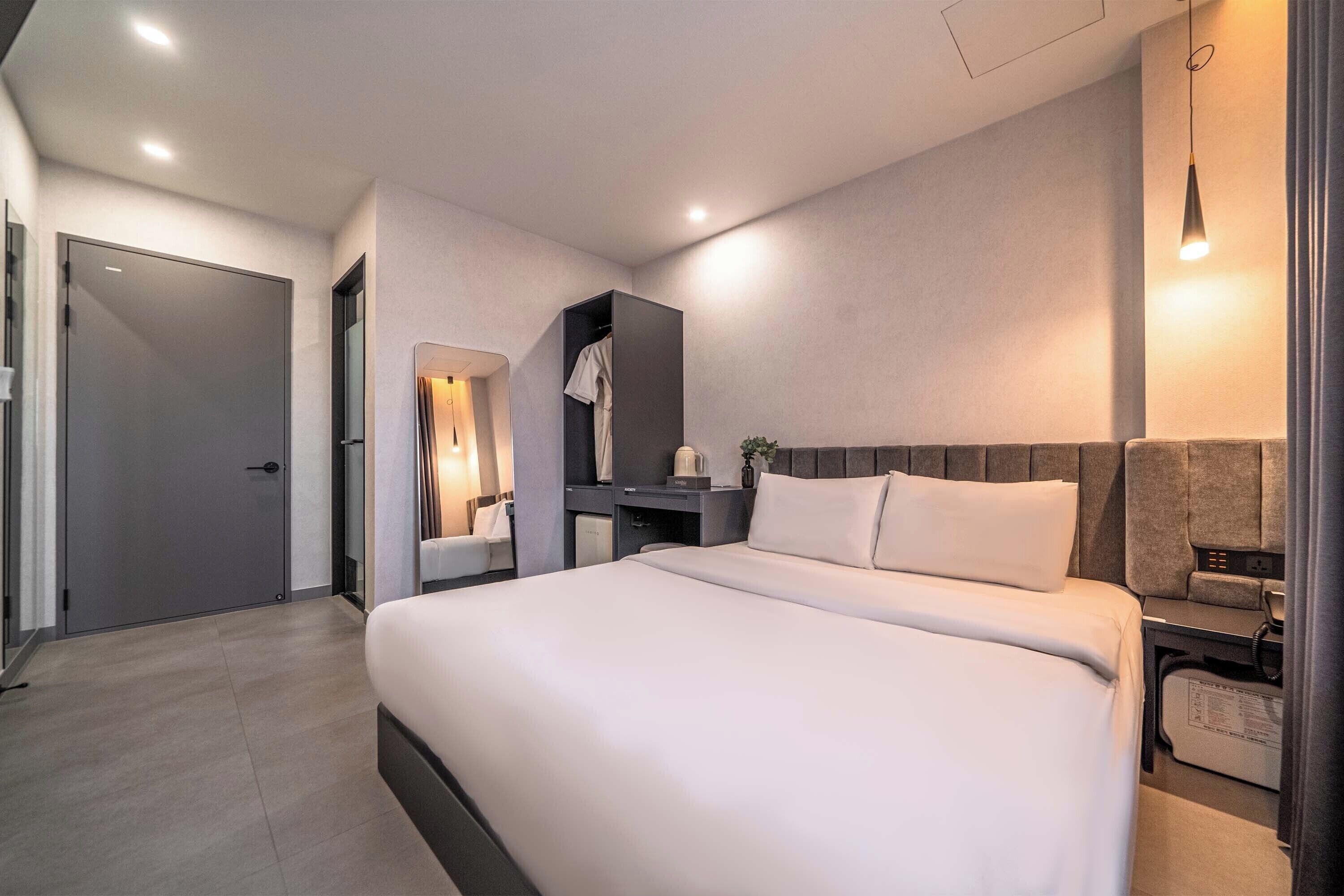 A modern hotel room with a large bed, bedside tables, a wardrobe, and a desk with a lamp.