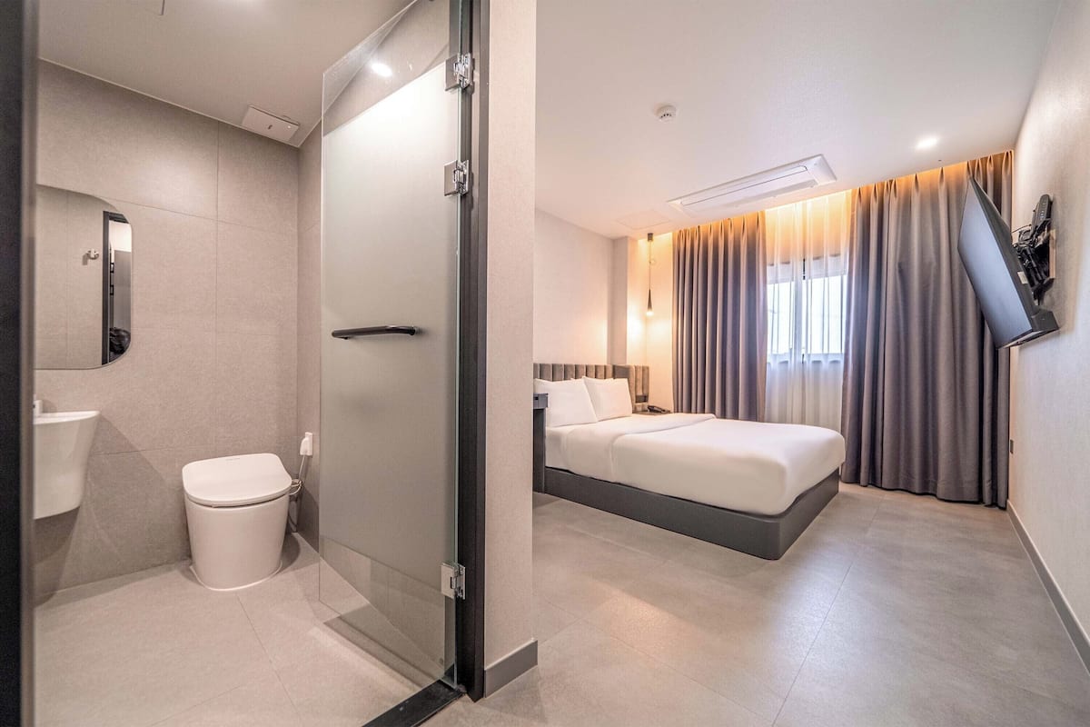 Habitación de hotel moderna con ducha, inodoro, cama y un amplio ventanal con cortinas.