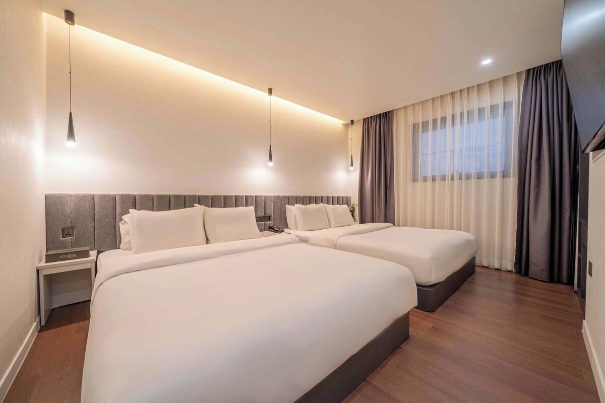 Habitación de hotel con dos camas, una mesita de noche, un televisor montado en la pared y una ventana con cortinas.