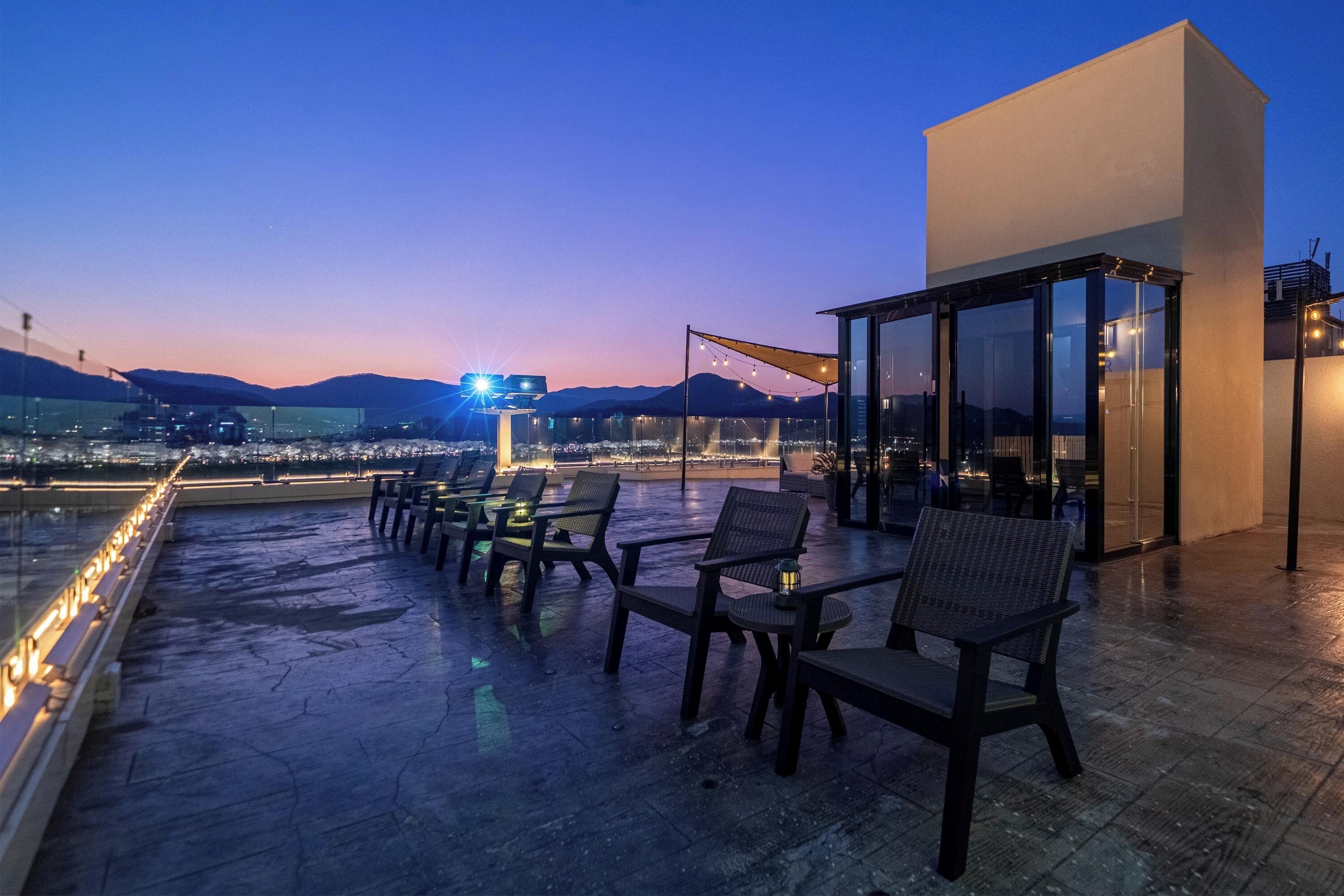 A rooftop terrace with chairs and tables, a glass enclosure, and a view of the cityscape at dusk.