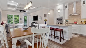 A spacious kitchen with white cabinetry, a dining area with a wooden table and white chairs, and a large island with stools. - Seashell Cove | Pet-friendly, Bikes, & Short walk to Egan's Creek! (Fernandina Beach)