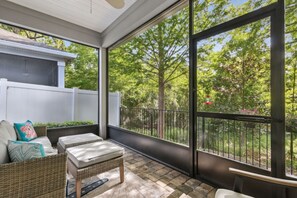 A balcony with wicker furniture, a white fence, and a view of trees and a garden. - Seashell Cove | Pet-friendly, Bikes, & Short walk to Egan's Creek! (Fernandina Beach)