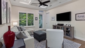 A living room with a sofa, ottoman, TV, and a ceiling fan. - Seashell Cove | Pet-friendly, Bikes, & Short walk to Egan's Creek! (Fernandina Beach)