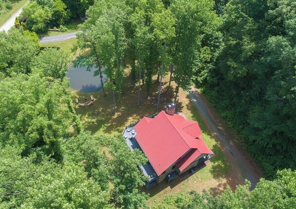 A single-story house with a red roof, surrounded by dense trees and a small pond.