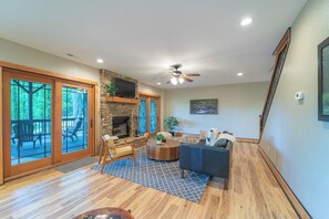 A spacious living room with a fireplace, a ceiling fan, and a large window overlooking a deck.