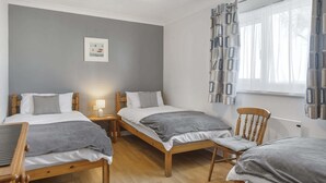 Quarto de hotel com duas camas de solteiro, uma cadeira de madeira, uma mesinha e uma janela com cortinas estampadas.