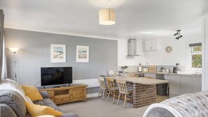 Uma sala de estar moderna com TV, sofás cinzas e um espaço de jantar com cadeiras de madeira e uma ilha de tijolos.