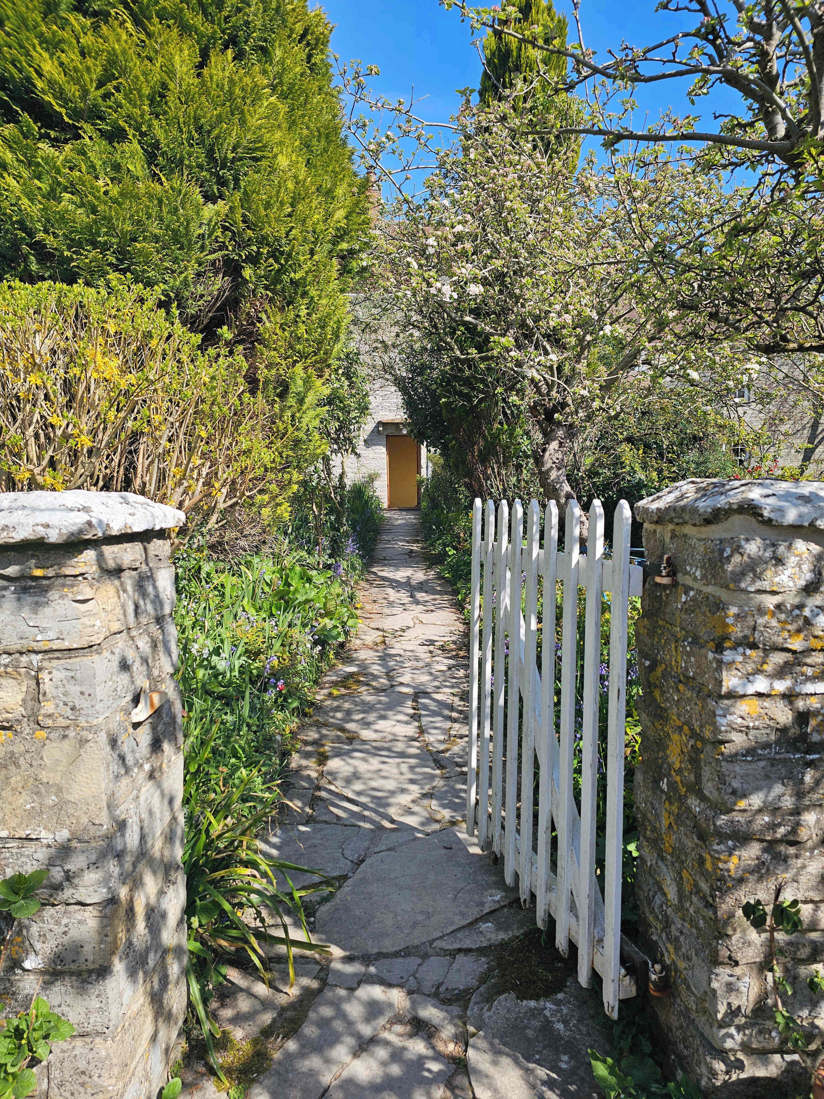 A stone pathway leading to a building with a yellow door, surrounded by lush greenery and a white picket fence.