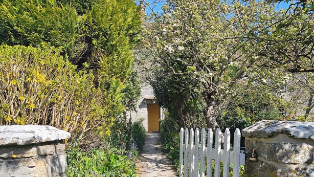 Un chemin de pierres mène à un bâtiment doté d’une porte jaune, entouré d’une végétation luxuriante et d’une clôture blanche en bois.