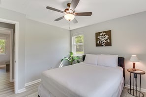 Een slaapkamer met een bed, een plafondventilator, een raam, een plant en een lamp.