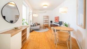 Un salon moderne avec un canapé gris, une table basse en bois et une table à manger ronde entourée de chaises. Un grand miroir orne le mur, ainsi qu’un lampadaire et un téléviseur posé sur un meuble.