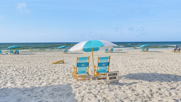 A beach with blue chairs and umbrellas, a sign indicating 'CUSTOMER WAITING HIGH WAVE'. - Balcony w/ Pool & Beach Views: Gulf Shores Condo! (Gulf Shores)