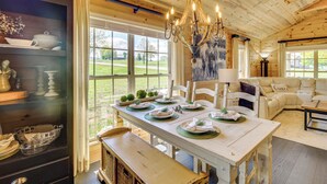 Een eetruimte met een houten tafel gedekt voor vier personen, een kroonluchter en uitzicht op het buitengebied door grote ramen.