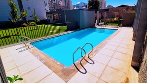 Piscina rectangular con agua azul cristalina, rodeada por una solana con baldosas y una cerca de metal.