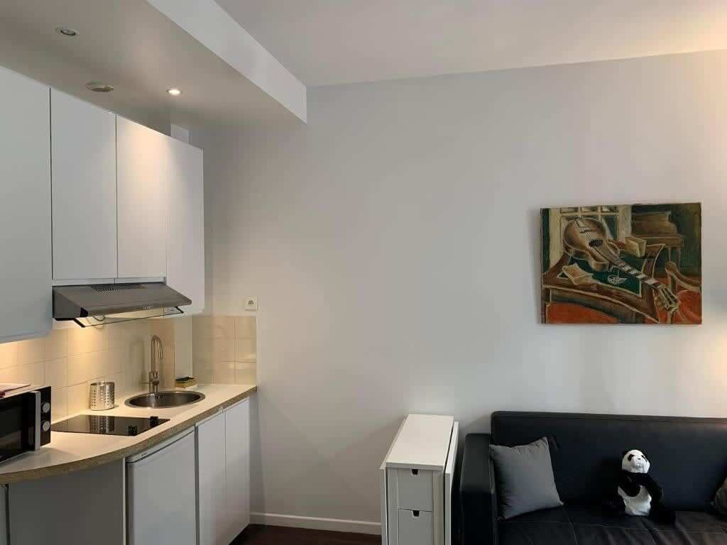 A modern kitchen with white cabinets, a microwave, and a sink. There is a black couch with a panda plush toy. A painting is on the wall.