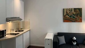 Una cocina moderna con armarios blancos, un microondas y un lavamanos. Hay un sofá negro con un peluche de panda. Un cuadro adorna la pared.