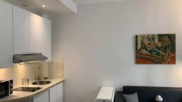 A modern kitchen with white cabinets, a microwave, and a sink. There is a black couch with a panda plush toy. A painting is on the wall.