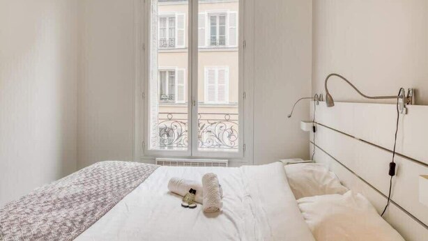 Habitación de hotel con cama, ventana con vistas a edificios y mesita de noche con lámpara.