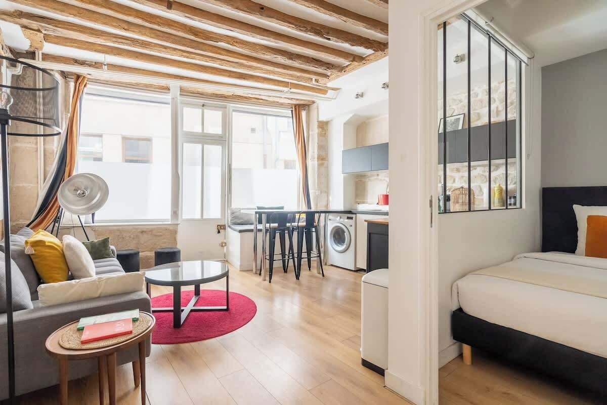 Un appartement moderne comprenant un salon, une cuisine et une chambre. Le salon est équipé d’un canapé, d’une table basse et d’un coin repas. La cuisine est dotée d’un lave-linge et d’une table à manger. La chambre comprend un lit et une fenêtre.