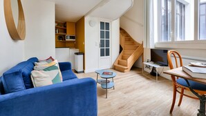 Een moderne woonkamer met een blauwe bank, een klein bijzettafeltje en een houten stoel. Een trap leidt naar een hoger niveau, en een raam met uitzicht op het omliggende landschap.