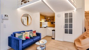 Een modern appartement met een blauwe bank, een klein rond tafeltje, een keukenhoek en een trap.