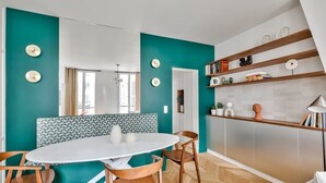 A modern kitchen with teal walls, a white dining table, wooden chairs, and a built-in bookshelf.