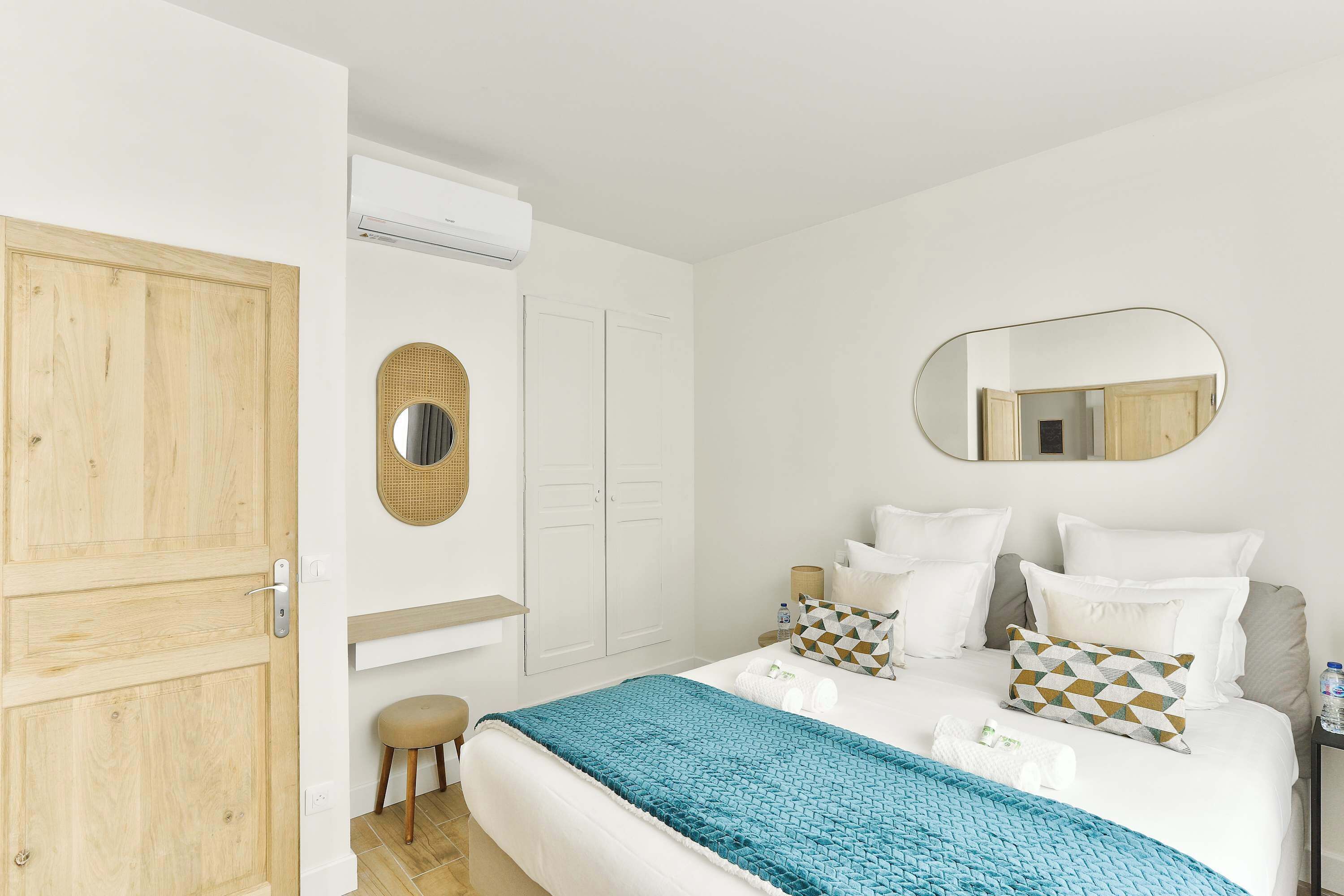 A bedroom with a bed, a wooden wardrobe, a round mirror, and a small stool.