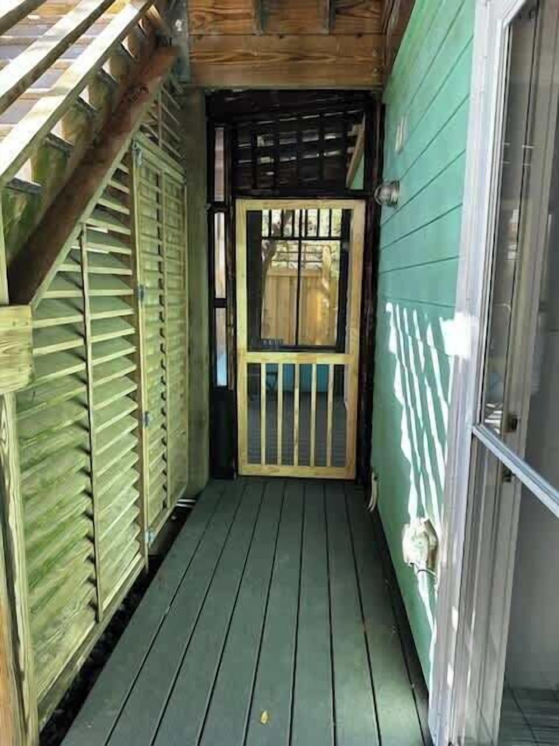 A narrow wooden deck leading to a door with a metal gate, a wooden staircase to the left, and a green wall on the right.