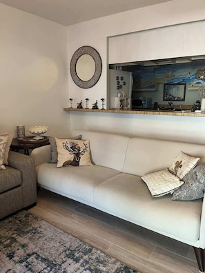Un salon comprenant un canapé, une table basse, un tapis, un miroir et un espace cuisine situé en mezzanine.