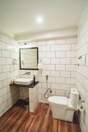A small, modern bathroom with white tiles, a wooden floor, a white sink, a mirror, and a toilet. - Dojee's Cottages (Srinagar)