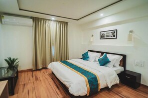 A hotel room with a wooden floor, a bed with white bedding and blue accents, a bedside table, a nightstand, a wall-mounted air conditioner, and a curtain. - Dojee's Cottages (Srinagar)