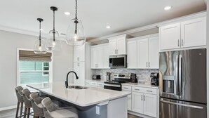 Cocina moderna con armarios blancos, electrodomésticos de acero inoxidable, una isla central con taburetes y luminarias colgantes, además de un ventanal con persianas.