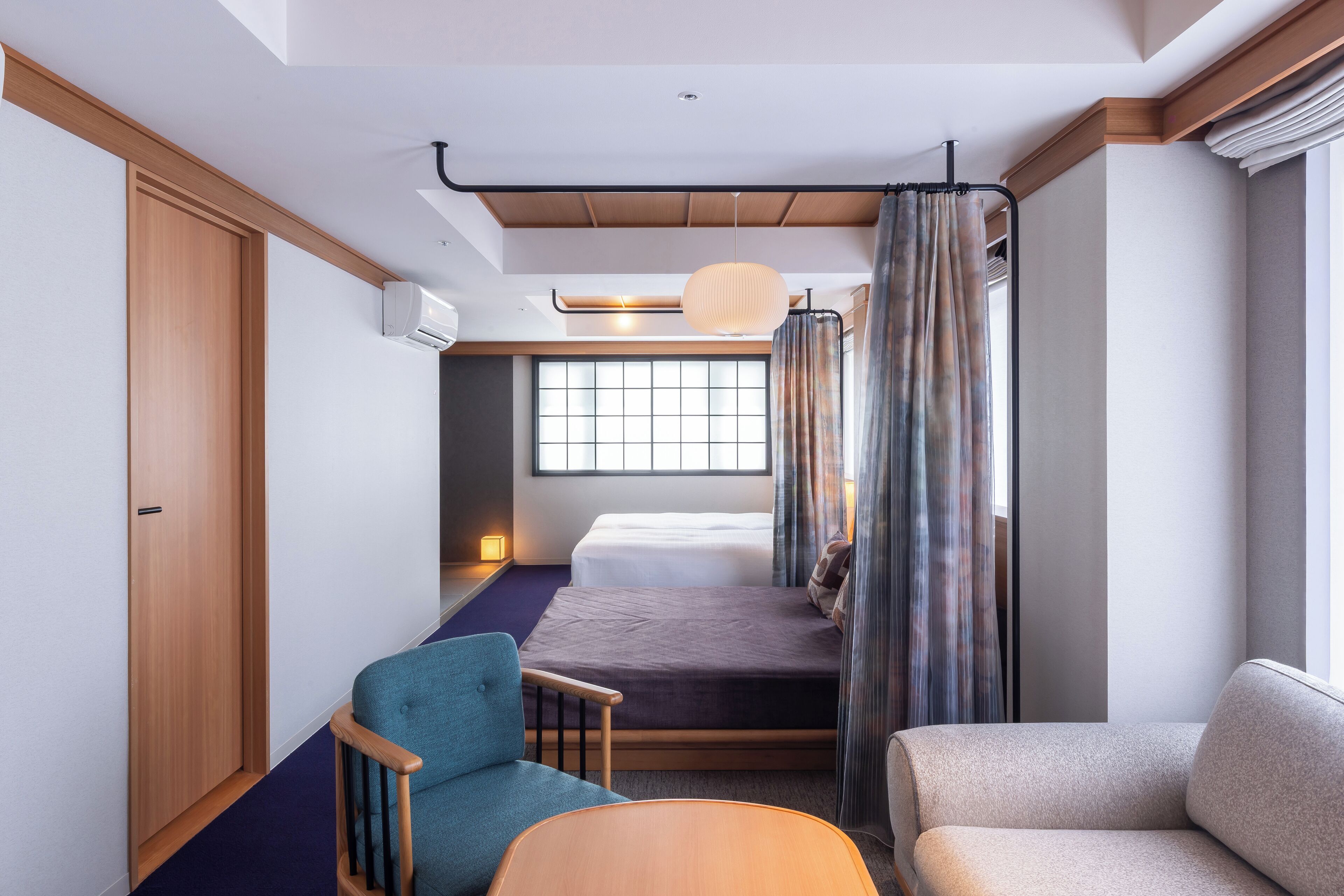 A compact hotel room with a bed, a seating area with a chair and sofa, a window with curtains, and a ceiling light.