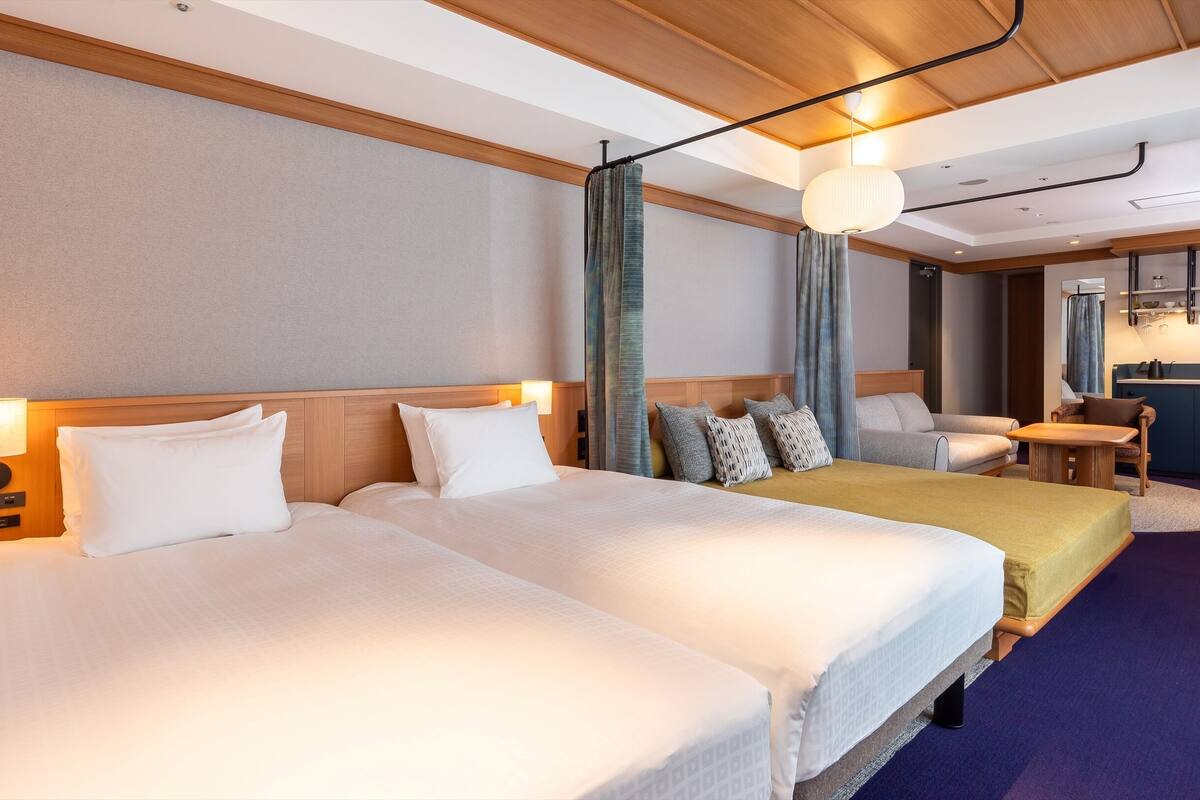 A hotel room with two beds, a sofa, a small table, a desk, and a kitchenette.