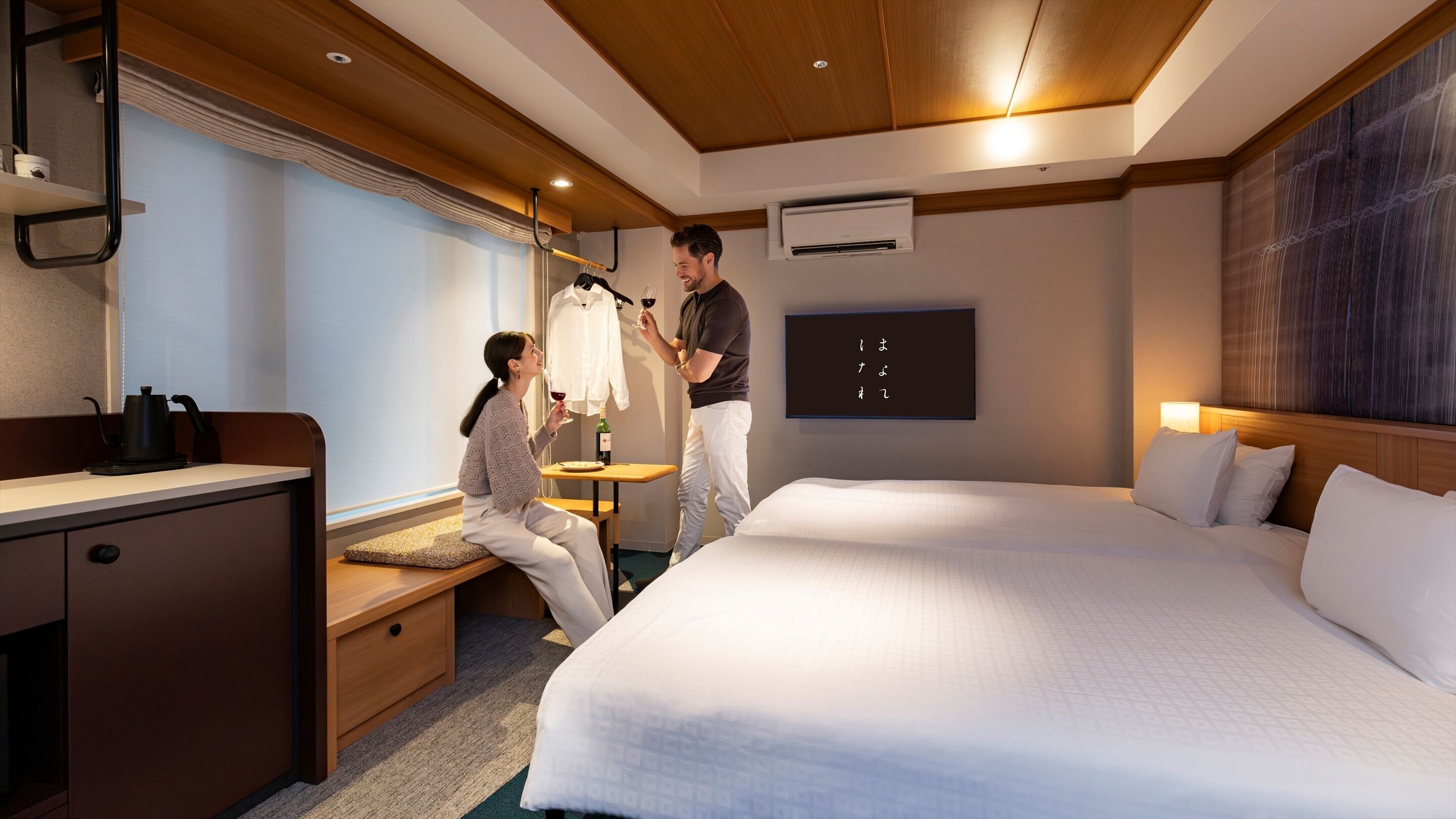 A hotel room with a bed, a desk, a chair, a window with curtains, an air conditioning unit, and a wall-mounted artwork.