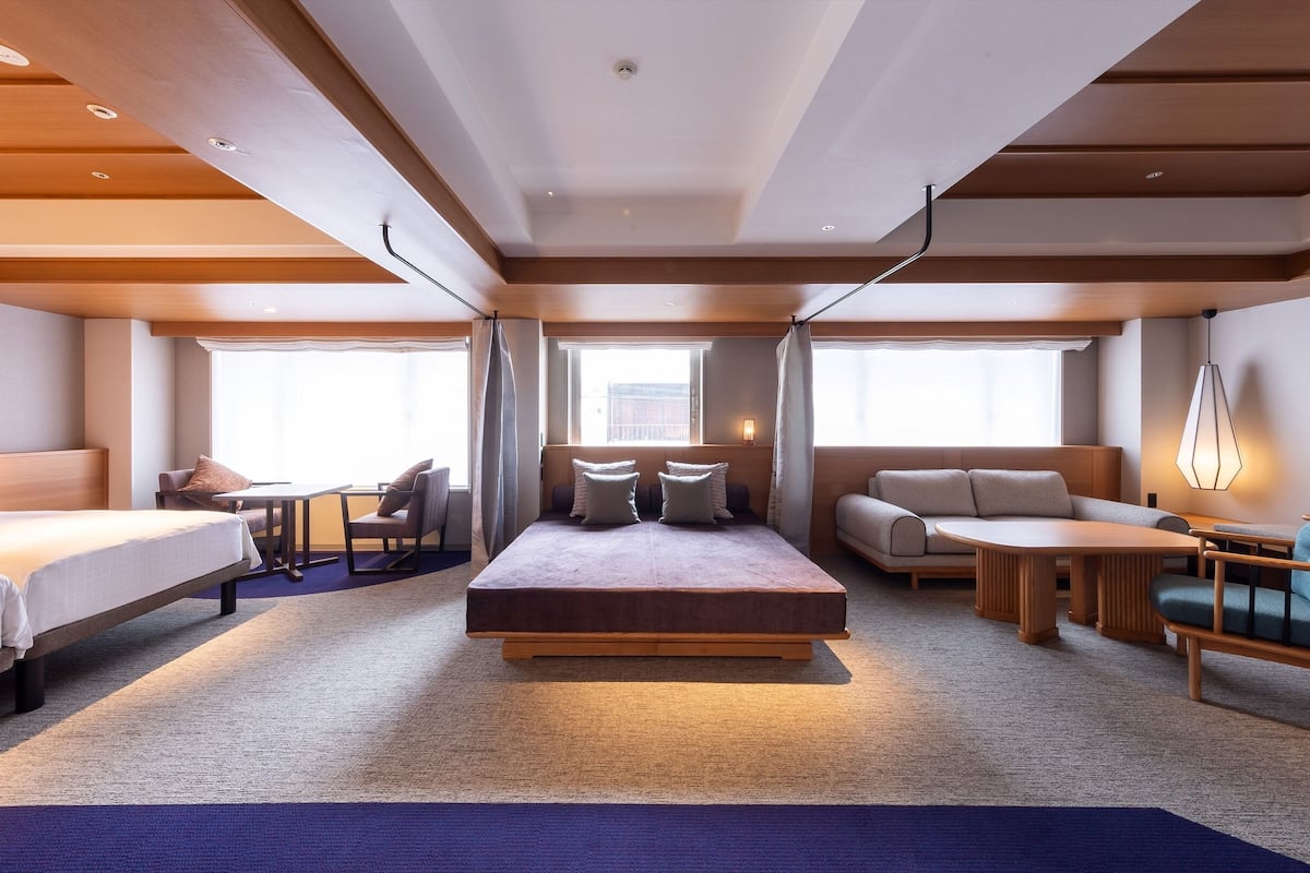 A hotel room with a bed, a sofa, a desk, and a chair. There are windows and a ceiling light.